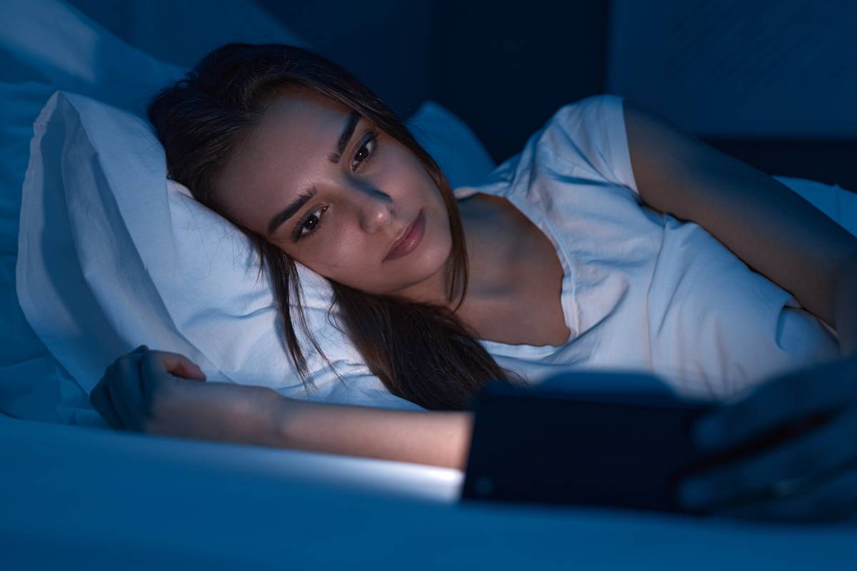 Female lying in bed and watching video on mobile phone in dark bedroom before sleep