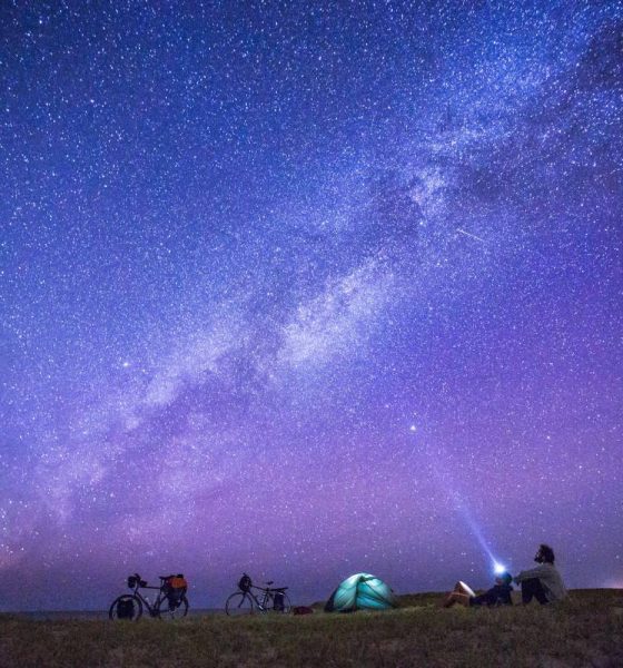 Anonymous tourists exploring numerous stars on sky in night while sitting on ground near camp tent.