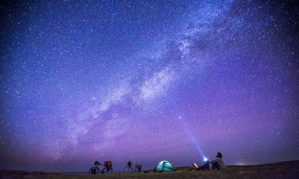 Anonymous tourists exploring numerous stars on sky in night while sitting on ground near camp tent.