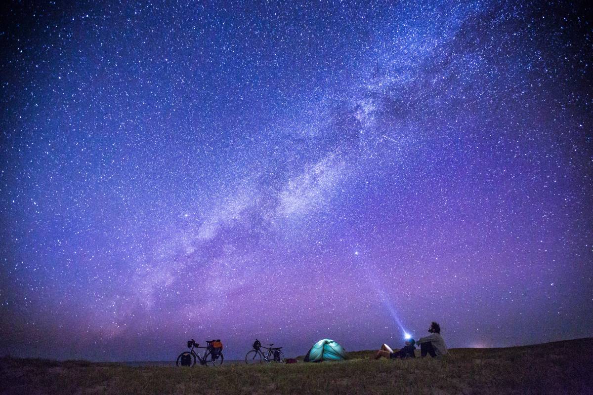 Anonymous tourists exploring numerous stars on sky in night while sitting on ground near camp tent.
